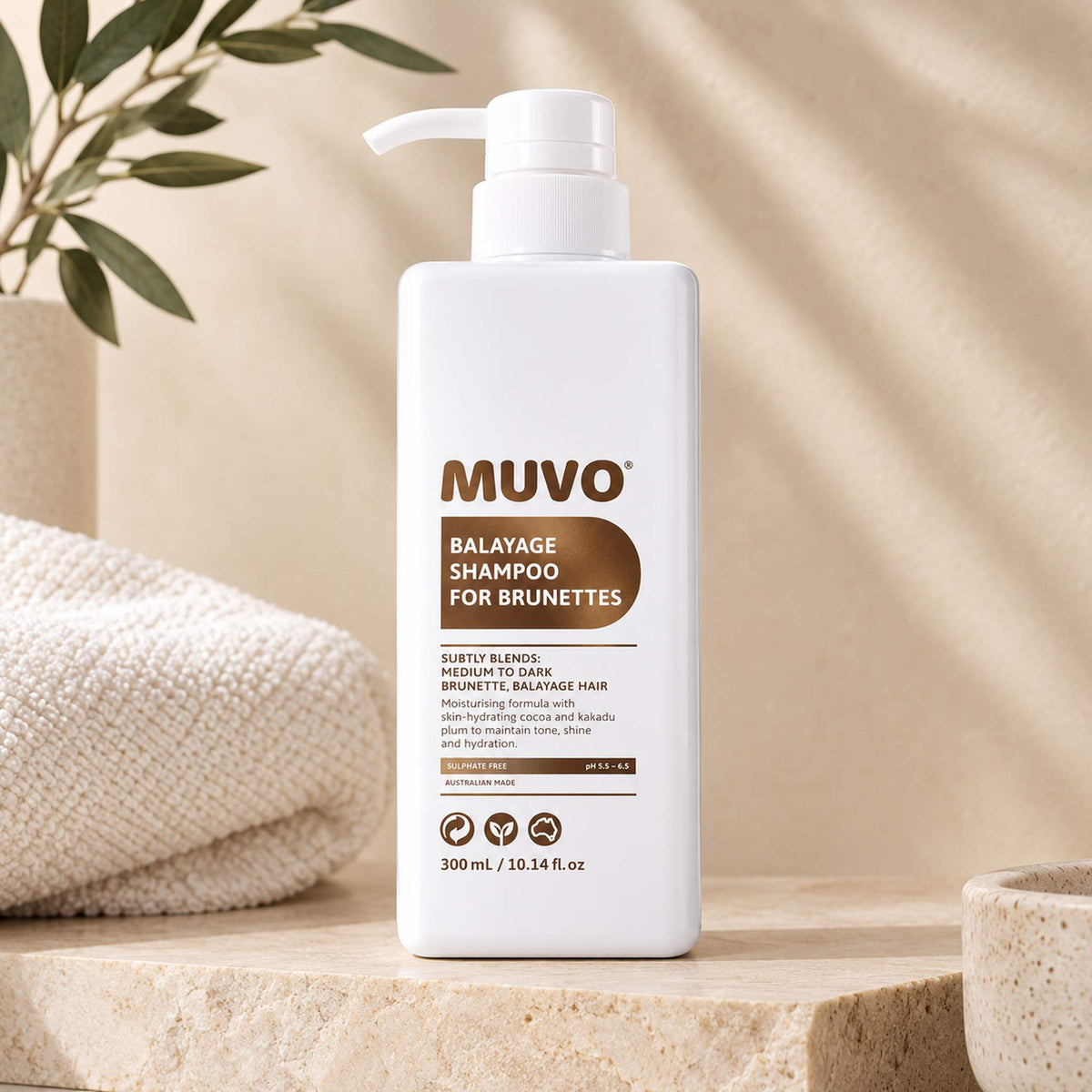 A white pump bottle of MUVO Balayage Shampoo for Brunettes 500ml, styled on a light stone surface with a folded cream towel, a potted plant, and soft beige drapery in the background.