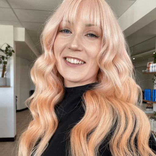 A smiling woman with long, wavy Peachy blonde hair and straight bangs, toned hair with Muvo Just Peachy Shampoo 300ml wearing a black top in an indoor setting.