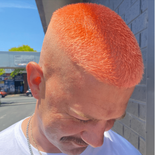 A close-up shot of a man outdoors with a shaved head with bright orange hair toned with Muvo Just Peachy Shampoo 300ml, wearing a white t-shirt and a silver chain necklace.