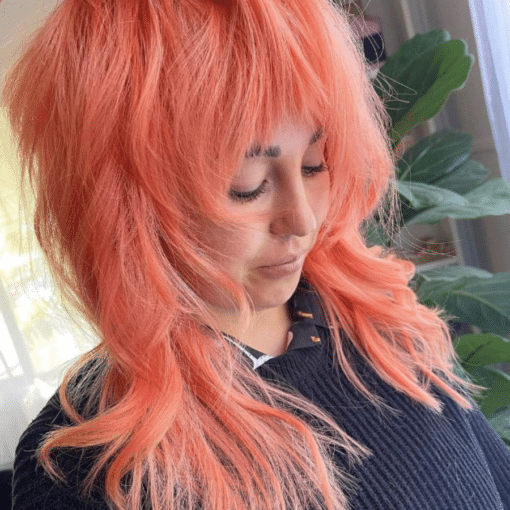 A close-up of a person with voluminous, layered wavy hair in a vibrant coral peach colour, toned with Muvo Just Peachy Shampoo 300ml wearing a dark ribbed knit top with a plant in the background.
